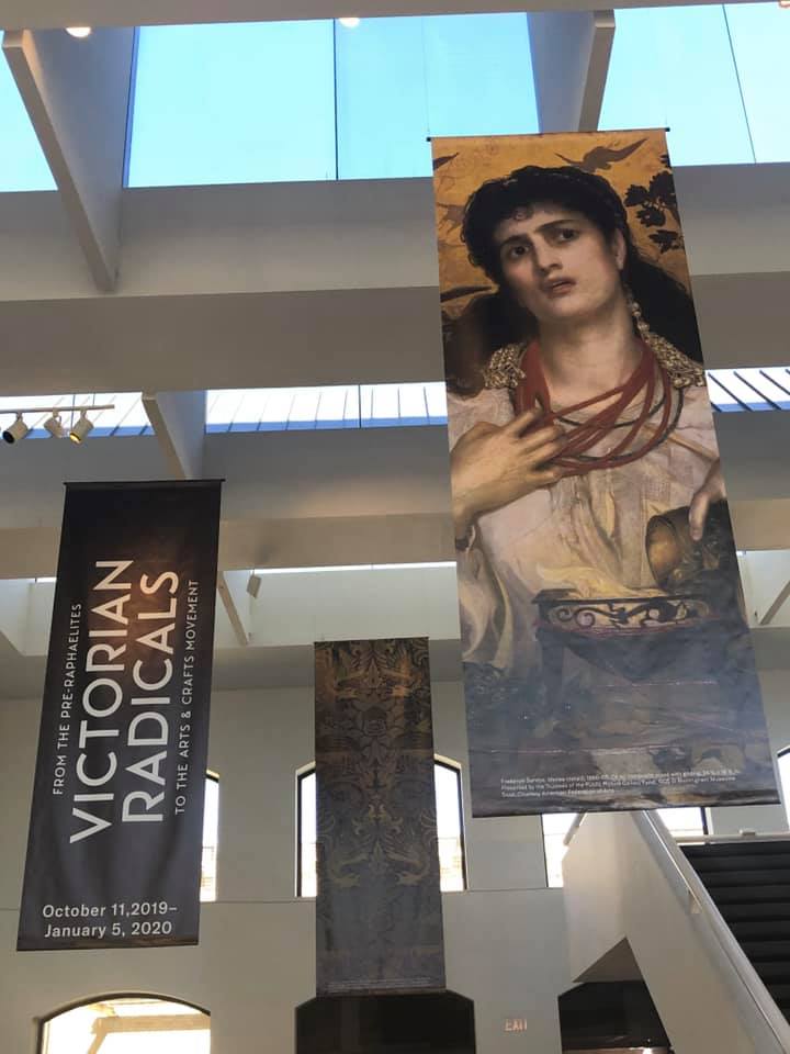 The image shows the interior of the San Antonio Museumn of Art with the exhibition banner for Victorian Radicals: from the Pre-Raphaelites to the Arts & Crafts Movement, as well as a banner showing the image of Medea by Pre-Raphaelite artist Frederick Sandys.